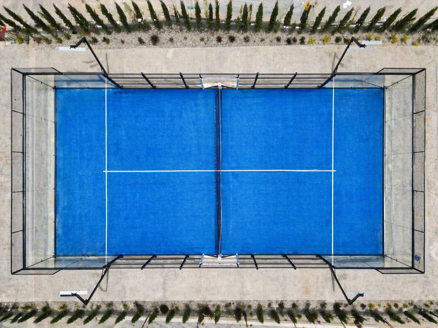 An aerial view of a rectangular pool with a blue surface, surrounded by a metal fence, and bordered by a concrete wall with vegetation.