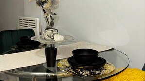 Een zithoek met een glazen tafel, een vaas met kunstbloemen, een zwarte kom en een geel pluizig kussen.