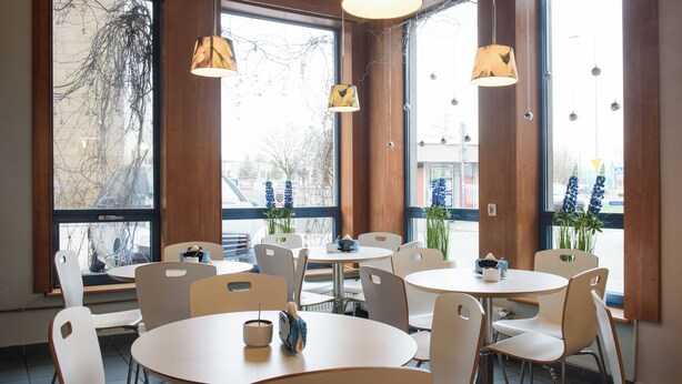 Un espace repas avec des tables rondes et des chaises blanches, de grandes fenêtres offrant une vue sur les arbres et les bâtiments, et des luminaires suspendus.