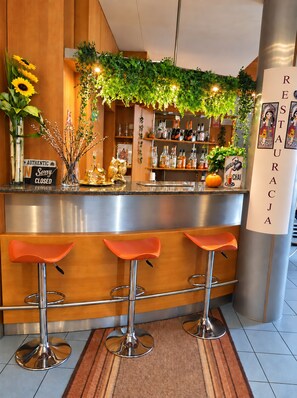 A bar area with a wooden counter, three orange bar stools, a sign indicating the bar is closed, and a wall with a green plant and a sign that reads 'RESTAURACION'.