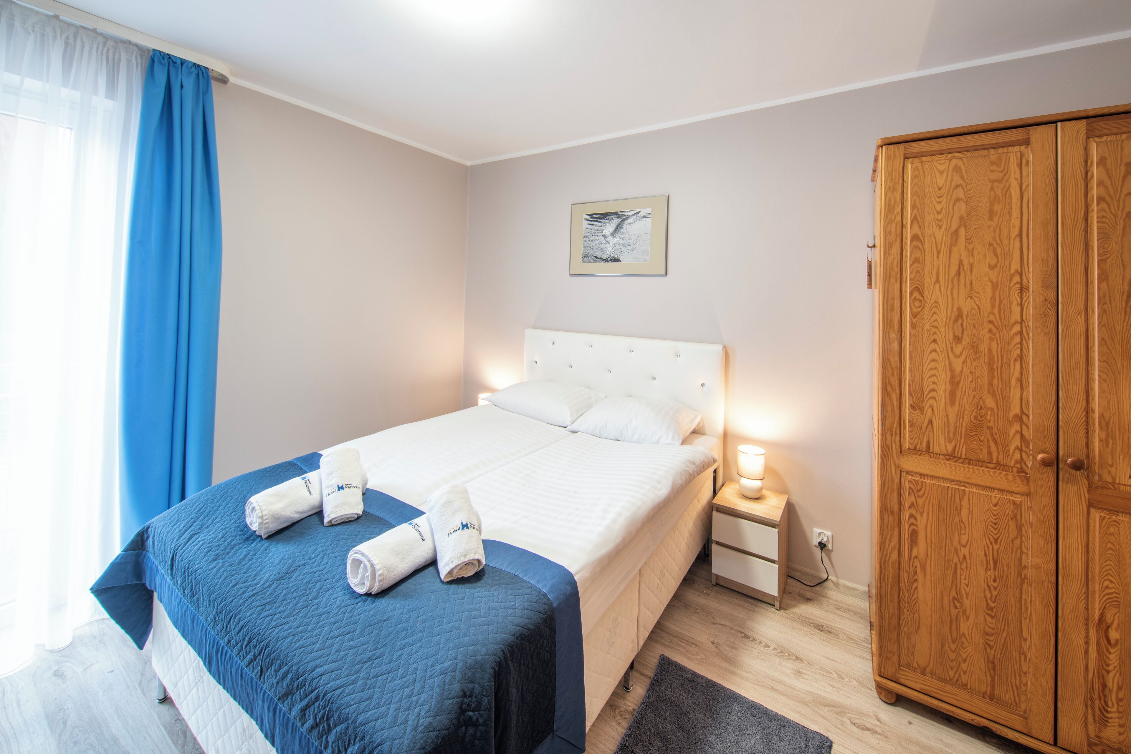 A neatly made bed with a blue bedspread and white pillows, a wooden wardrobe, a bedside table with a lamp, and a framed picture on the wall.