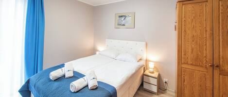 A neatly made bed with a blue bedspread and white pillows, a wooden wardrobe, a bedside table with a lamp, and a framed picture on the wall.