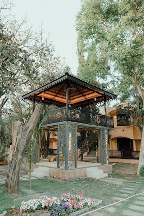 A gazebo with a metal roof and decorative panels, surrounded by trees and a flower bed.
