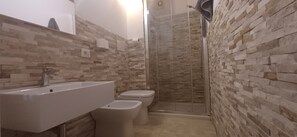 A bathroom with a stone-patterned wall, a sink, two toilets, and a glass-enclosed shower. - Super Central Rooms (Cagliari)
