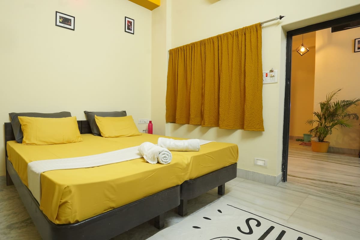A hotel room with a bed, two pillows, a white towel, a yellow curtain, and a potted plant.