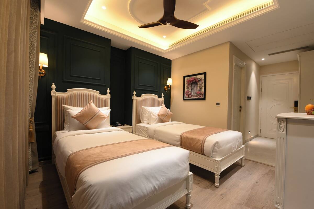 A hotel room with two beds, a ceiling fan, a painting, and a small kitchenette.