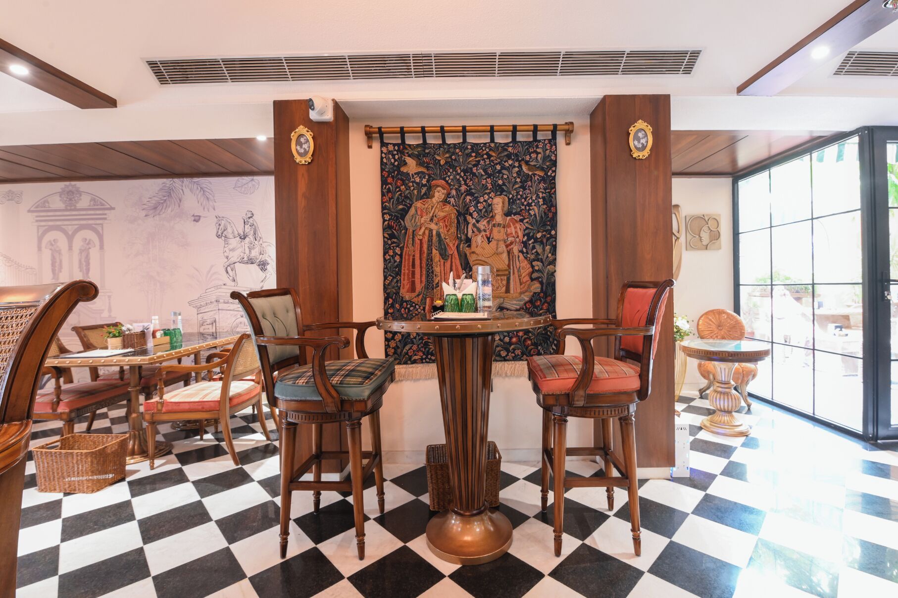 Un hall d’hôtel avec un sol à carreaux, des meubles en bois, une grande tapisserie au centre et une grande fenêtre.