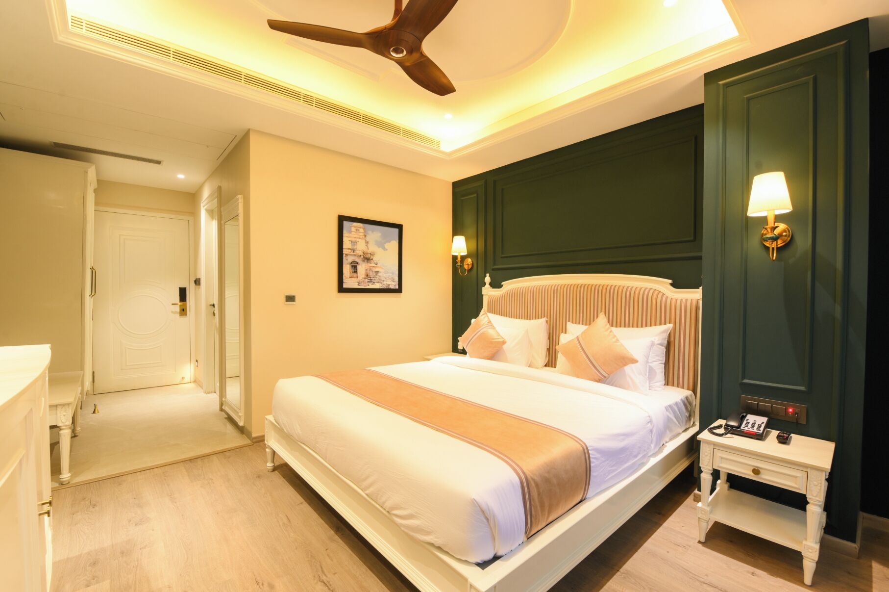 Une chambre d’hôtel avec un grand lit, une table de chevet avec une lampe, un ventilateur de plafond et une porte donnant sur une autre pièce.