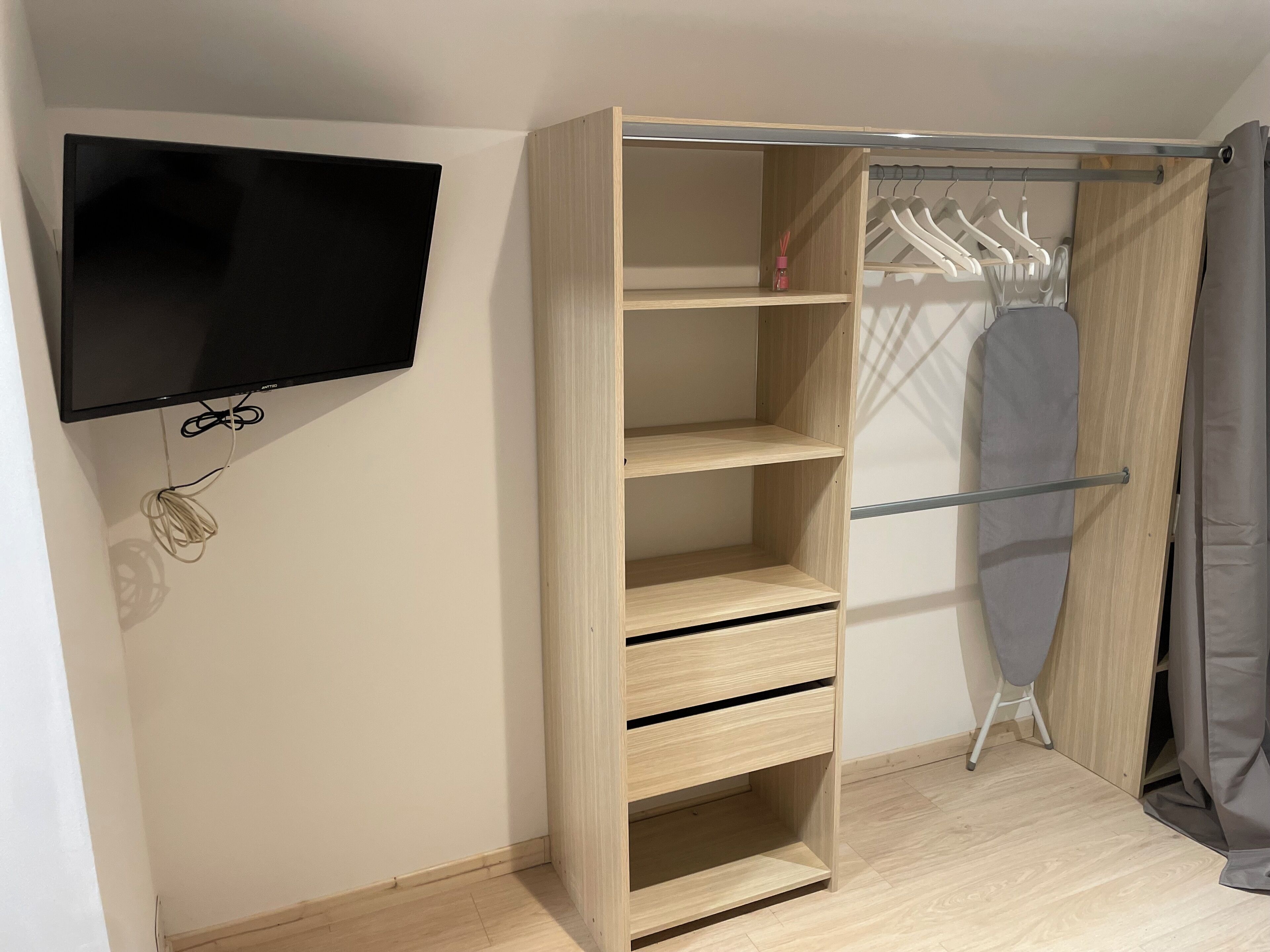 A room with a built-in wooden wardrobe, a flat-screen TV mounted on the wall, and a clothing rack with hangers.