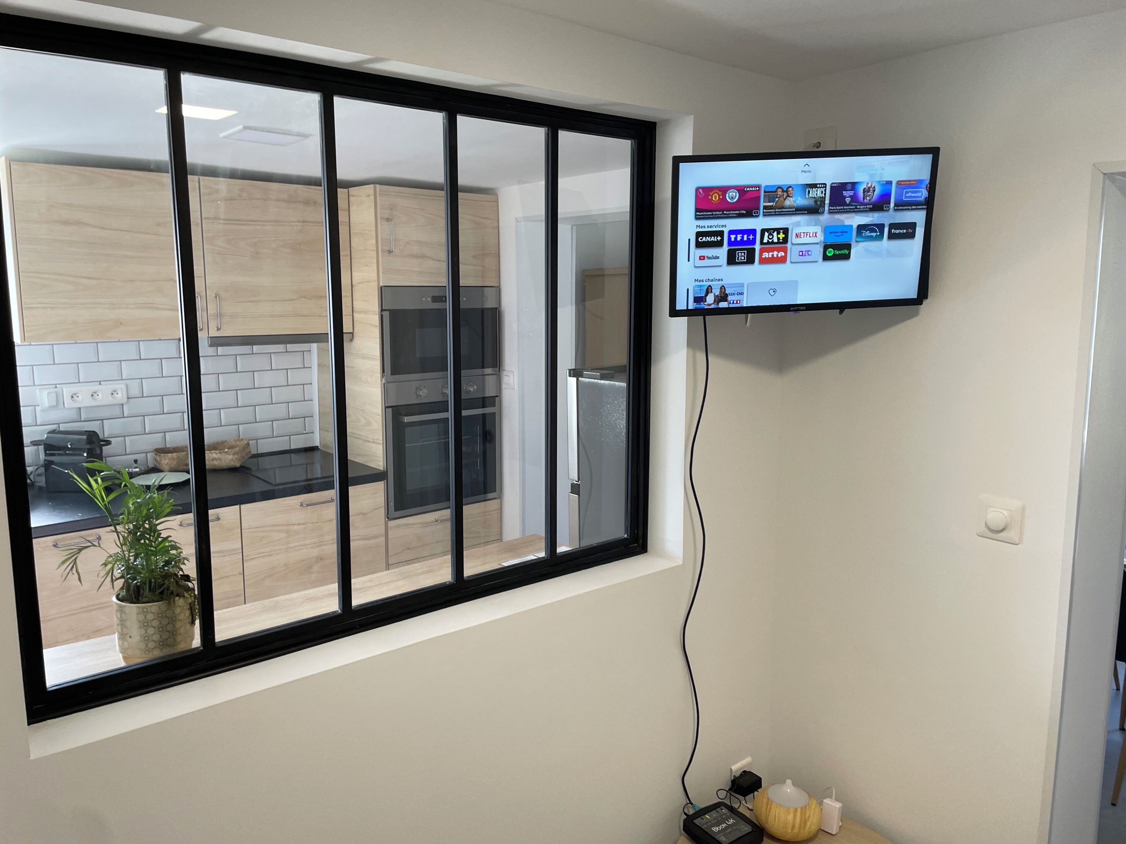 A modern kitchen with a large window, a flat-screen TV mounted on the wall, and a power strip on the floor.