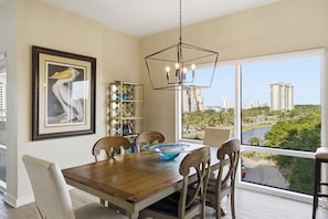 A dining area with a wooden table, chairs, a chandelier, a framed artwork, and a view of a cityscape through large windows. - 1BR Luau on 5th Floor - spanning views of the lagoon pool and Gulf at Sandestin Golf & Beach Resort (Miramar Beach)