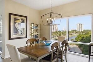 A dining area with a wooden table, chairs, a chandelier, a framed artwork, and a view of a cityscape through large windows.