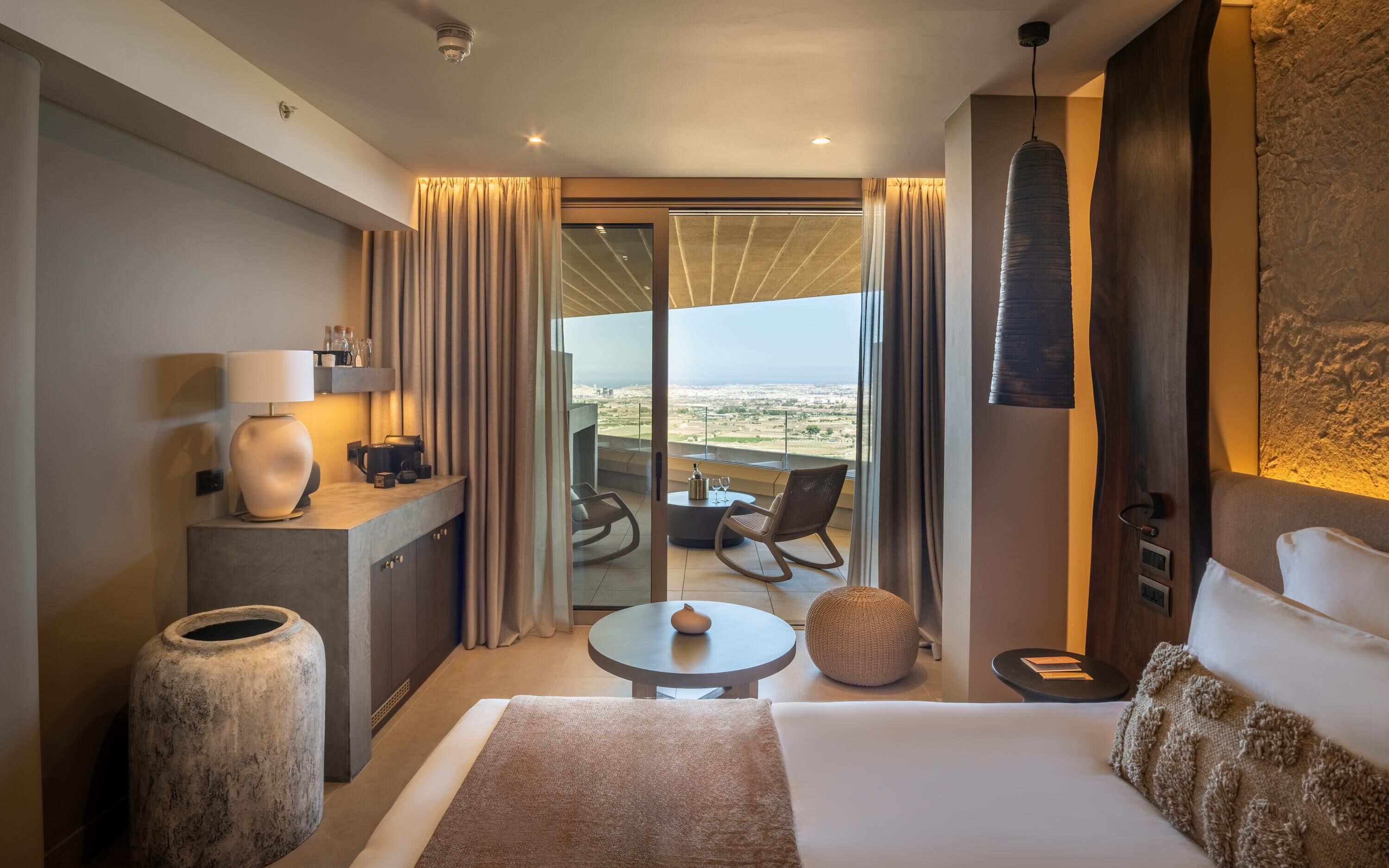 A modern hotel room with a large bed, a small table, a chair, a sofa, a large vase, a lamp, and a view of the city through the balcony.