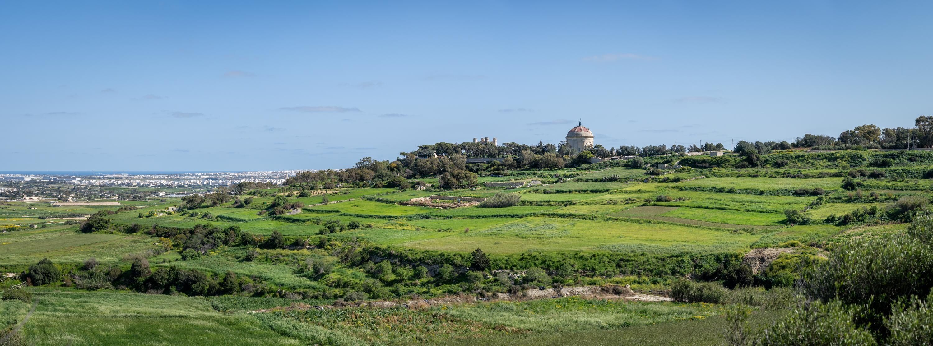 A hilltop view of a historic building with a dome, surrounded by terraced fields and a distant cityscape.