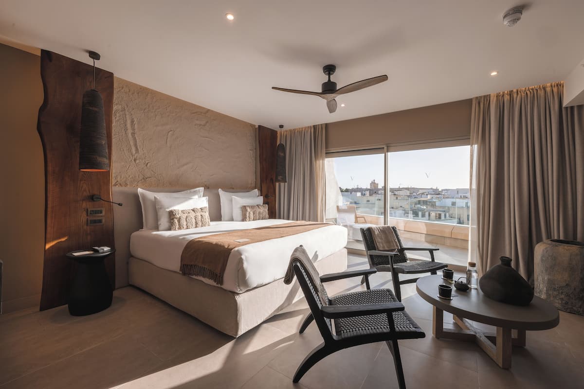 Une chambre d’hôtel moderne dotée d’un grand lit, d’un coin salon avec une table et des chaises, d’une vue sur la ville grâce à de grandes fenêtres et d’un ventilateur de plafond.