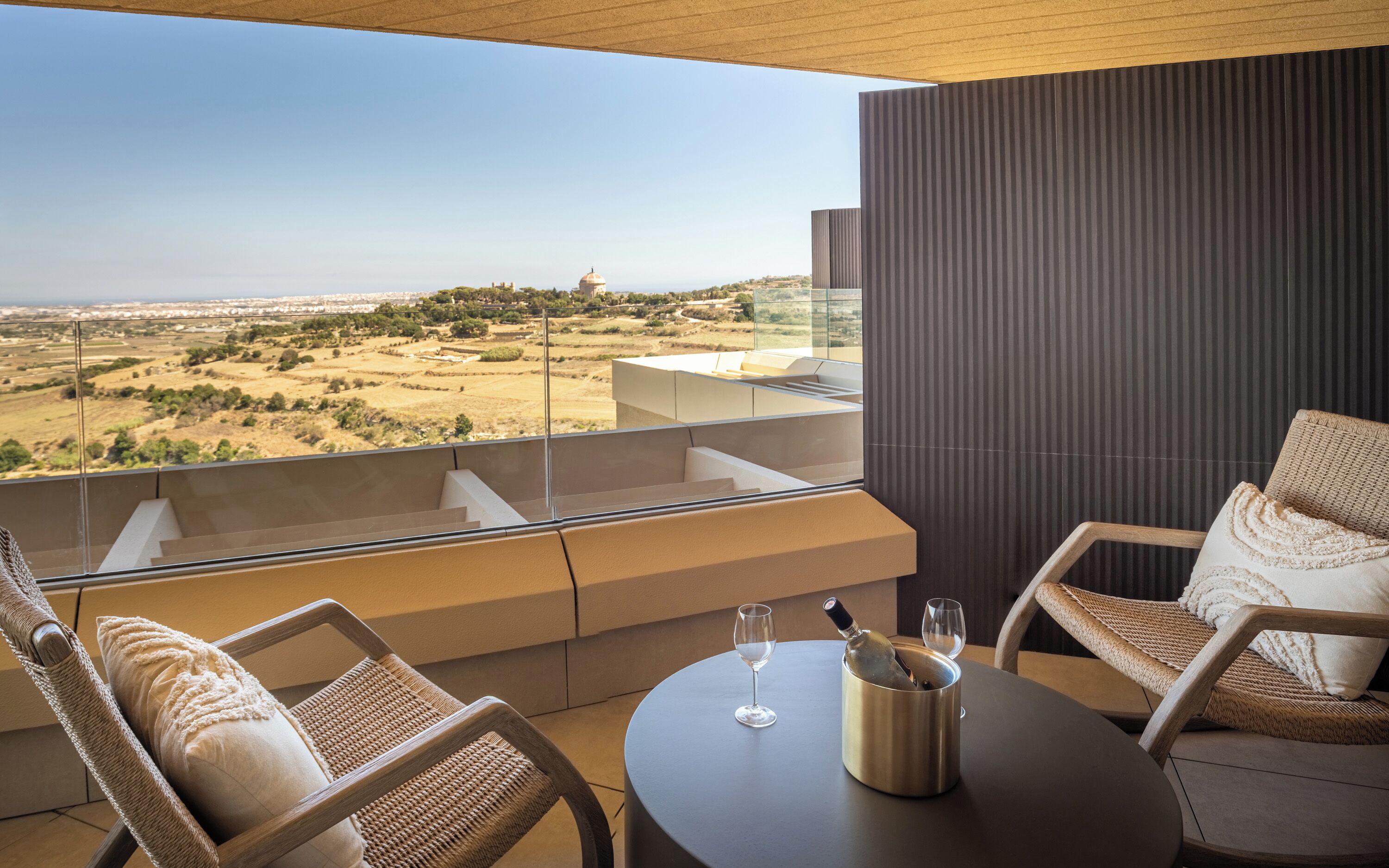 A balcony with a view of a hilly landscape, two wicker chairs, a table with a wine glass and a bottle, and a dark textured wall.