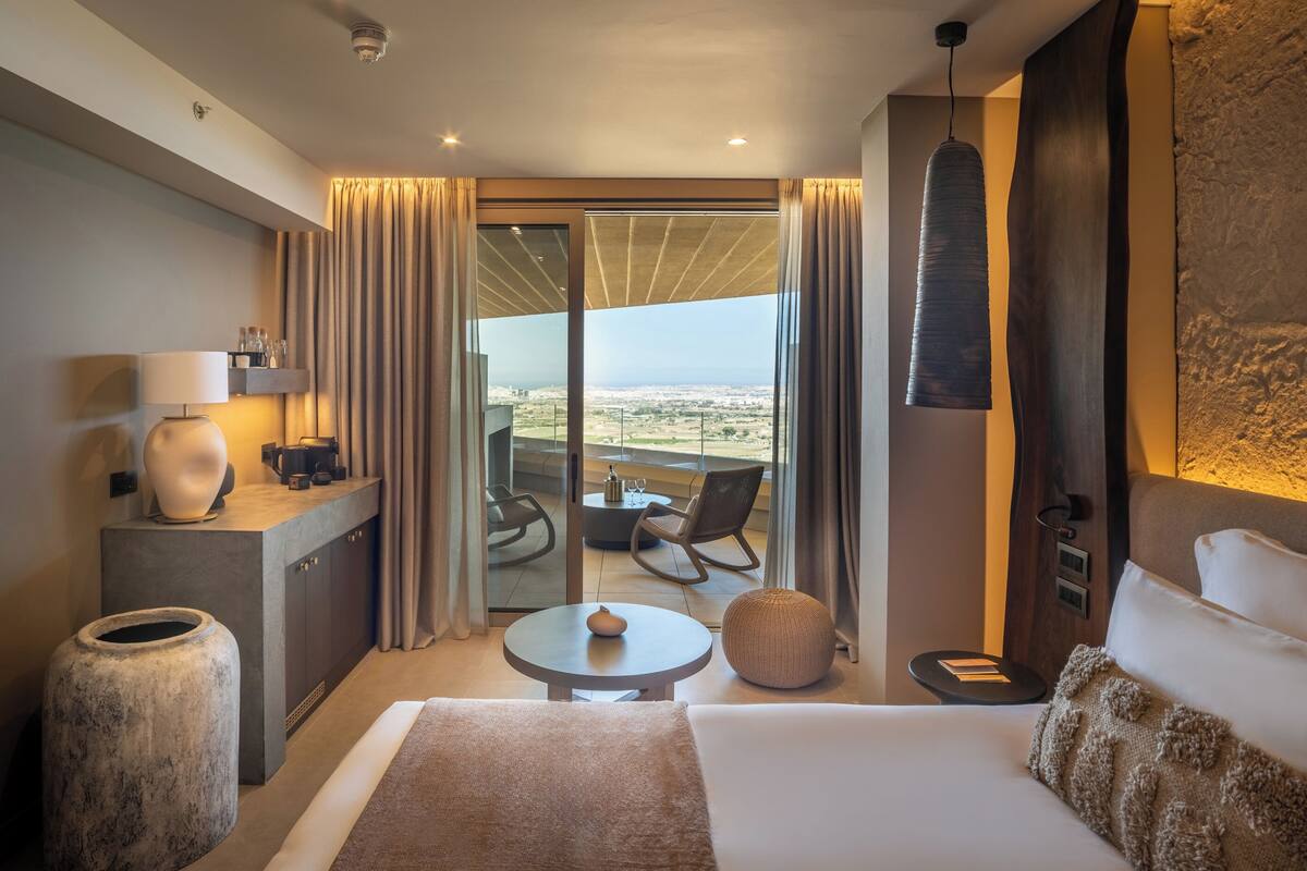 Une chambre d’hôtel moderne avec un grand lit, une petite table ronde, un canapé, une vue sur la ville et un balcon équipé d’une table et de chaises.