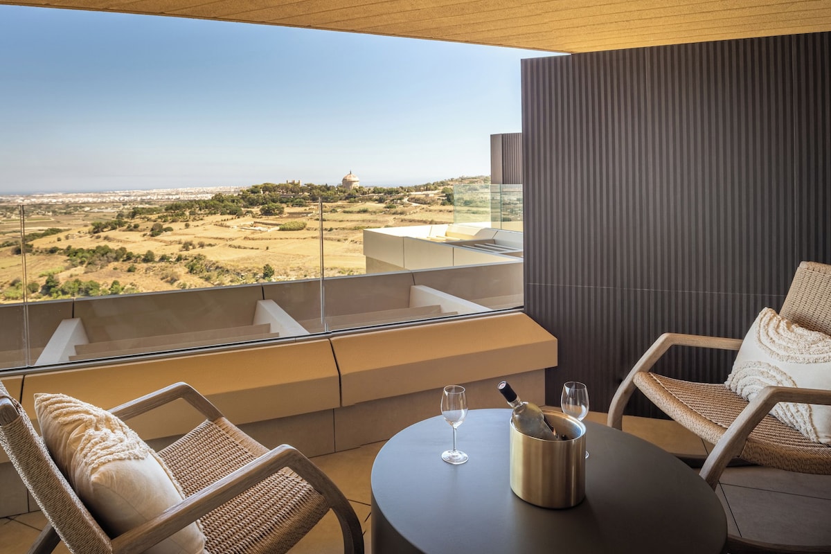 A balcony with a view of a hilly landscape, two wicker chairs, a table with a wine glass and a bottle, and a dark textured wall.
