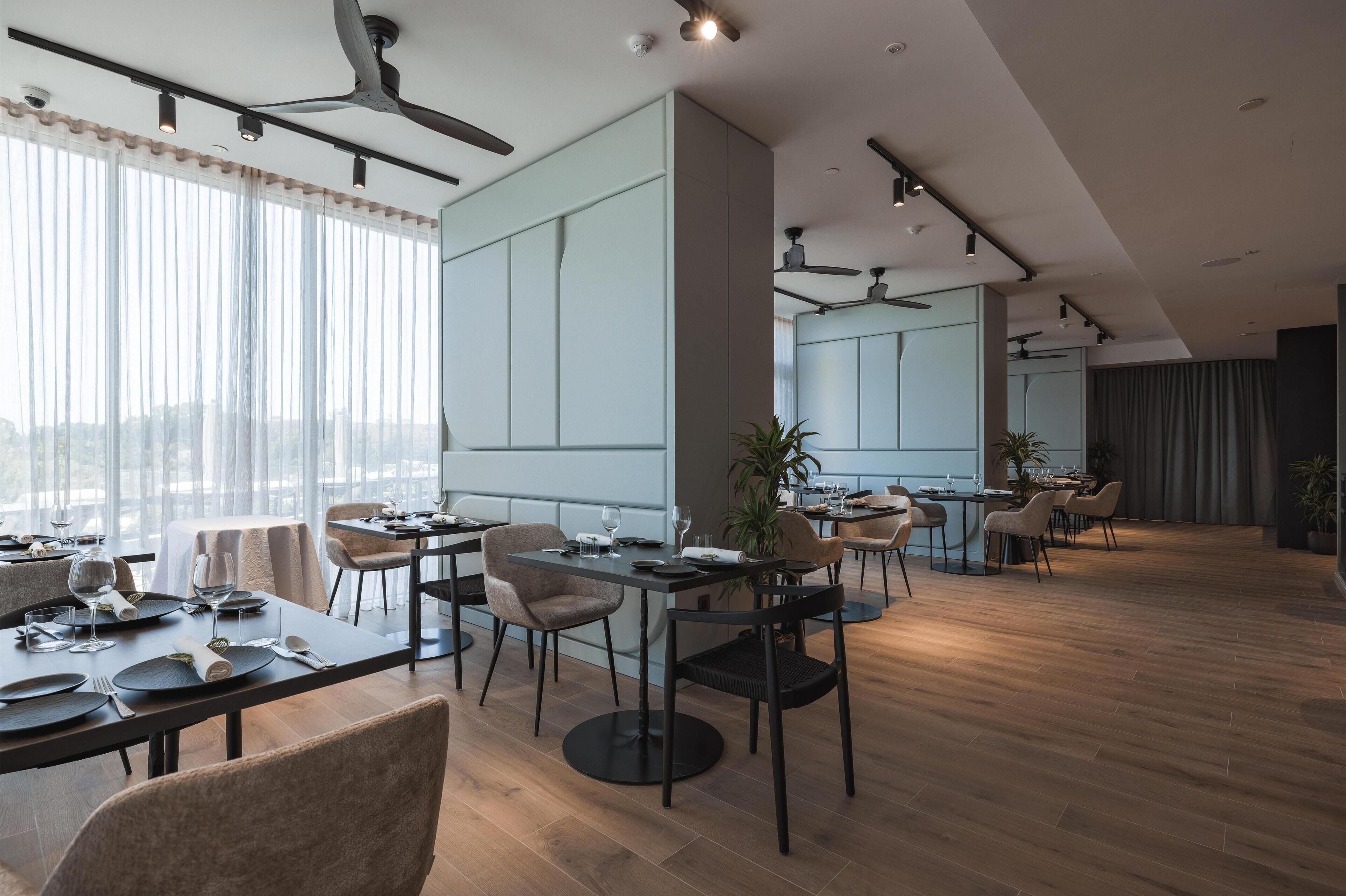 A modern dining area with tables set for a meal, a kitchen in the background, and a view of the city through large windows.