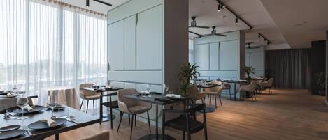A modern dining area with tables set for a meal, a kitchen in the background, and a view of the city through large windows.
