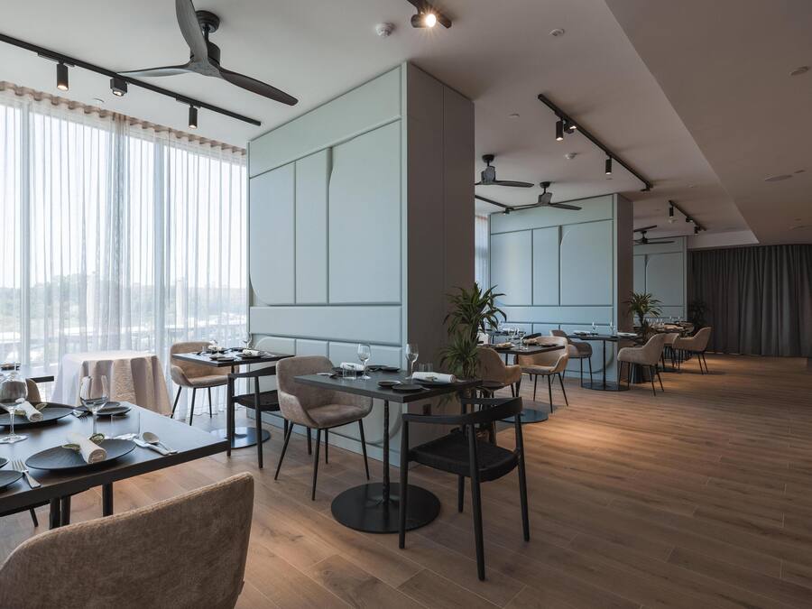 Un comedor moderno con mesas dispuestas para una comida, una cocina al fondo y vistas de la ciudad a través de grandes ventanales.