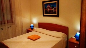 Una cama bien hecha con una colcha blanca y un almohadón naranja, dos mesitas de noche con lámparas, un cuadro enmarcado en la pared y una ventana con cortinas traslúcidas.