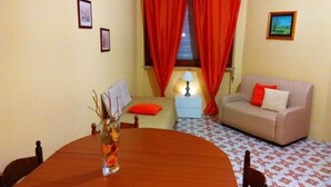 Un comedor con mesa de madera, sillas, un jarrón con ramas, un sofá con cojines rojos, una mesita con lámpara y una ventana con cortinas rojas.