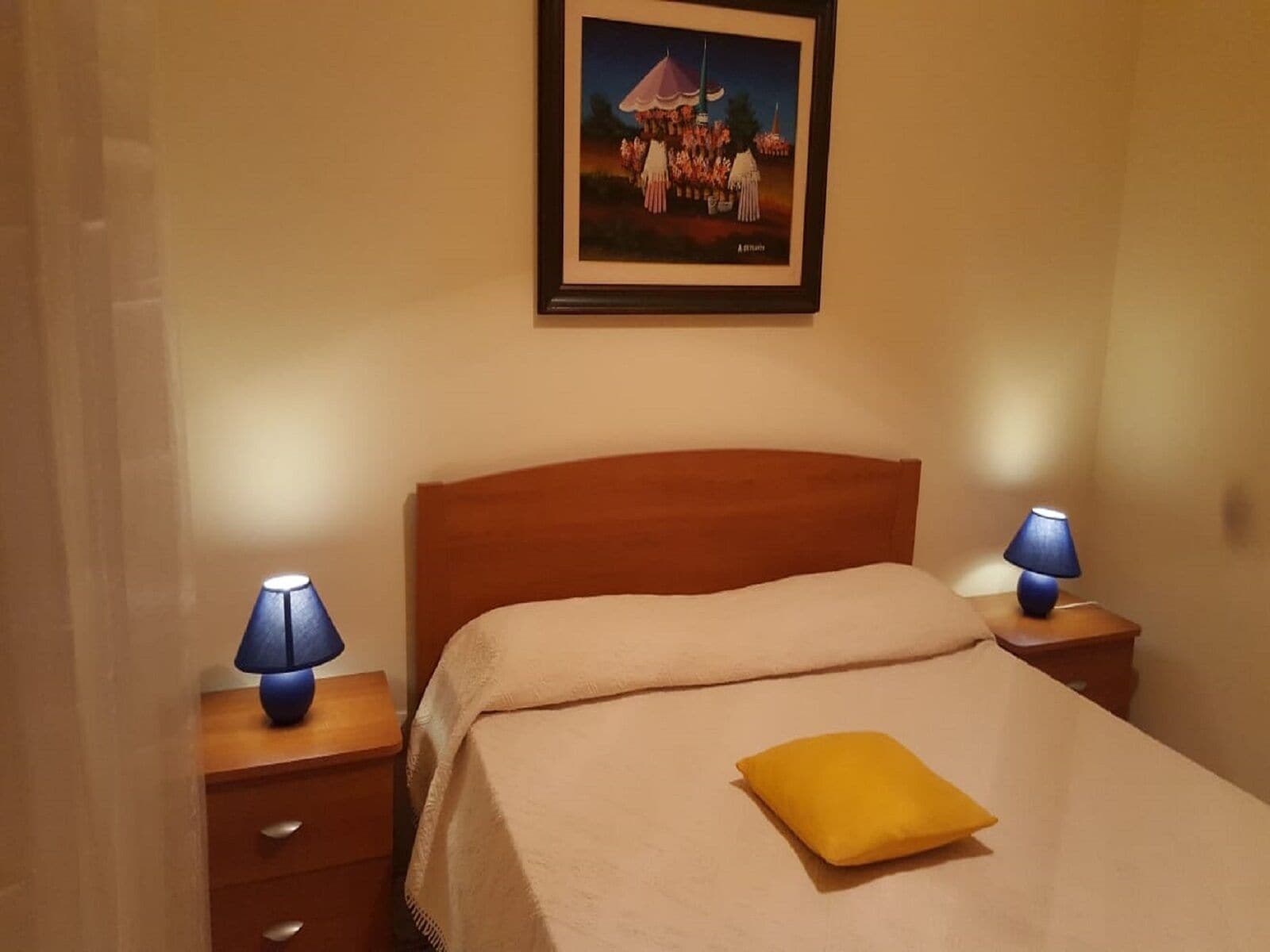 A bedroom with a wooden headboard, a bed with a beige blanket and a yellow pillow, two bedside tables with lamps, and a framed picture on the wall.