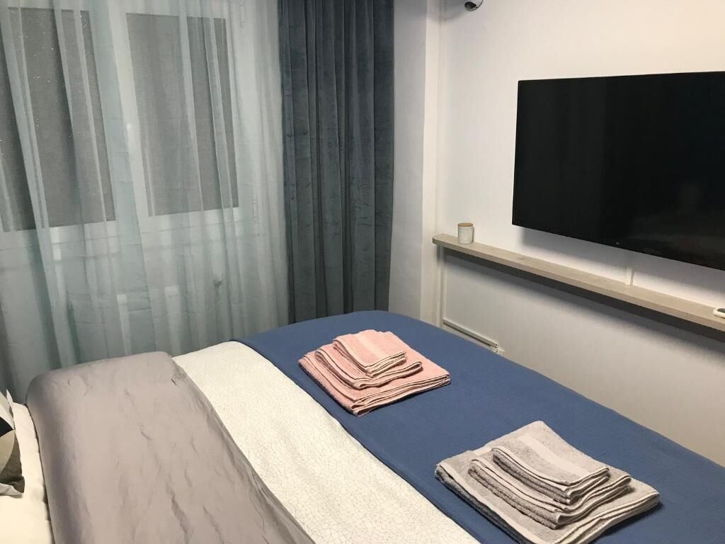 A hotel room with a bed, a flat-screen TV mounted on the wall, and a window with sheer curtains.