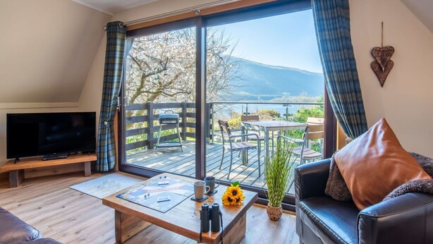 A living room with a TV, a coffee table with a board game, a sofa, and a view of a deck with outdoor seating. - Wonderful private villa for 4 guests with WIFI, TV, balcony and panoramic view (Kilfinnan)