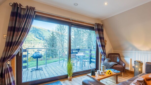 A cozy living room with a large window, a leather sofa, a wooden coffee table, and a view of a deck with outdoor furniture. - Stunning private villa for 4 guests with WIFI, TV, balcony and panoramic view (Kilfinnan)