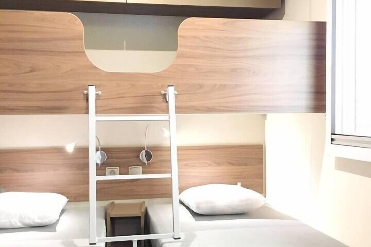 A compact room with two beds, a small table, and a shelf unit.