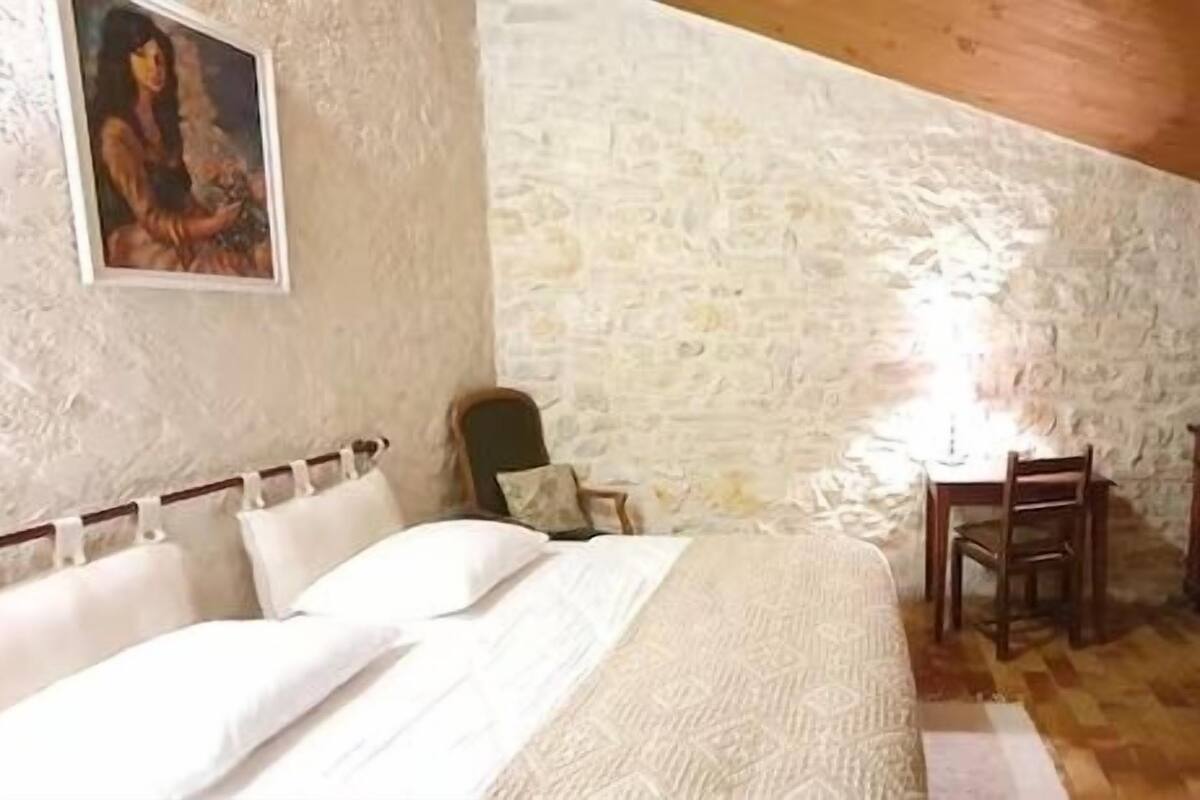 A bedroom with a stone wall, a bed with white bedding, a wooden dresser, and a chair.