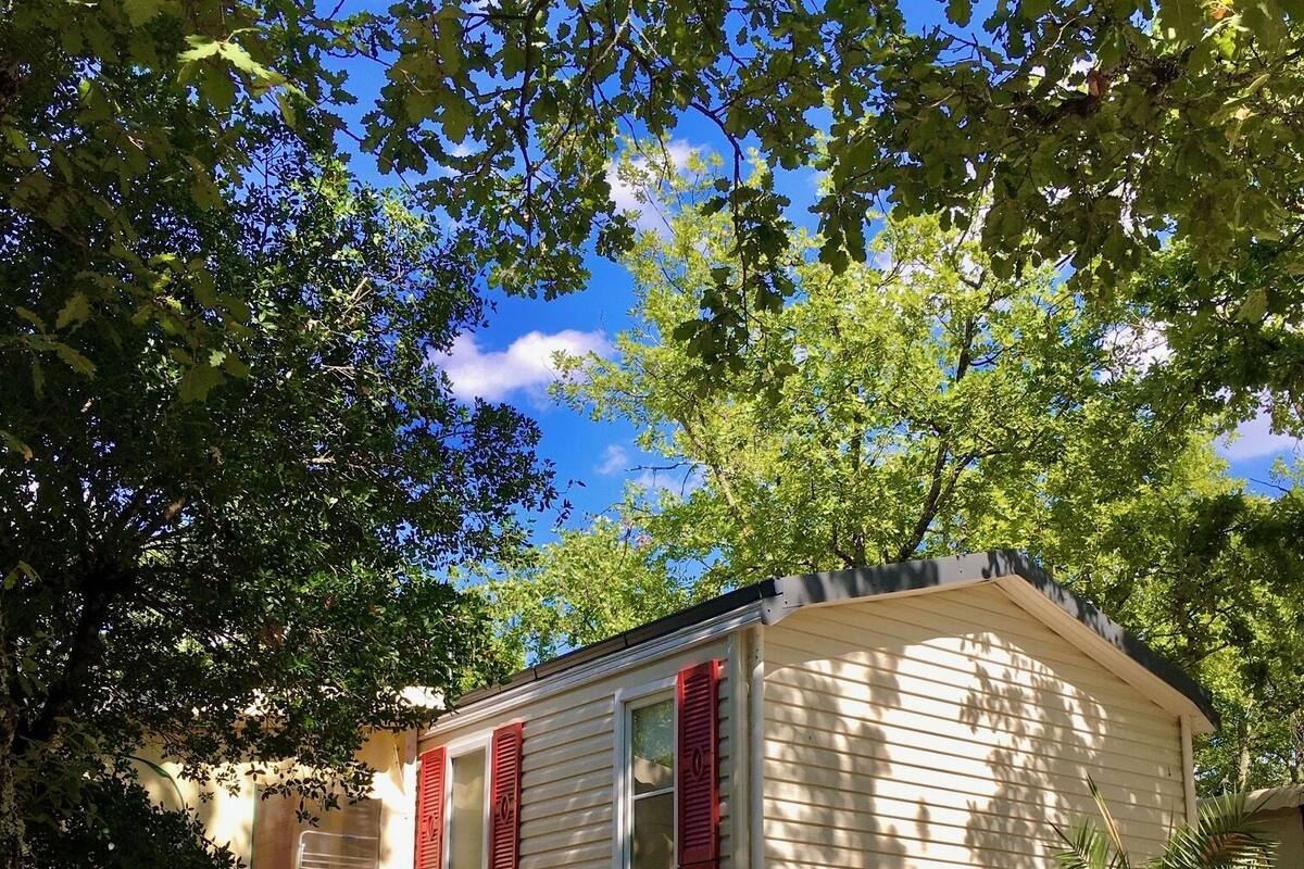 A small, single-story mobile home with red shutters, surrounded by trees and a gravel driveway.
