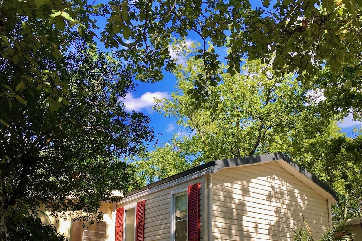 Une petite maison mobile d’un seul étage, avec des volets rouges, entourée d’arbres et bordée d’une allée en gravier.