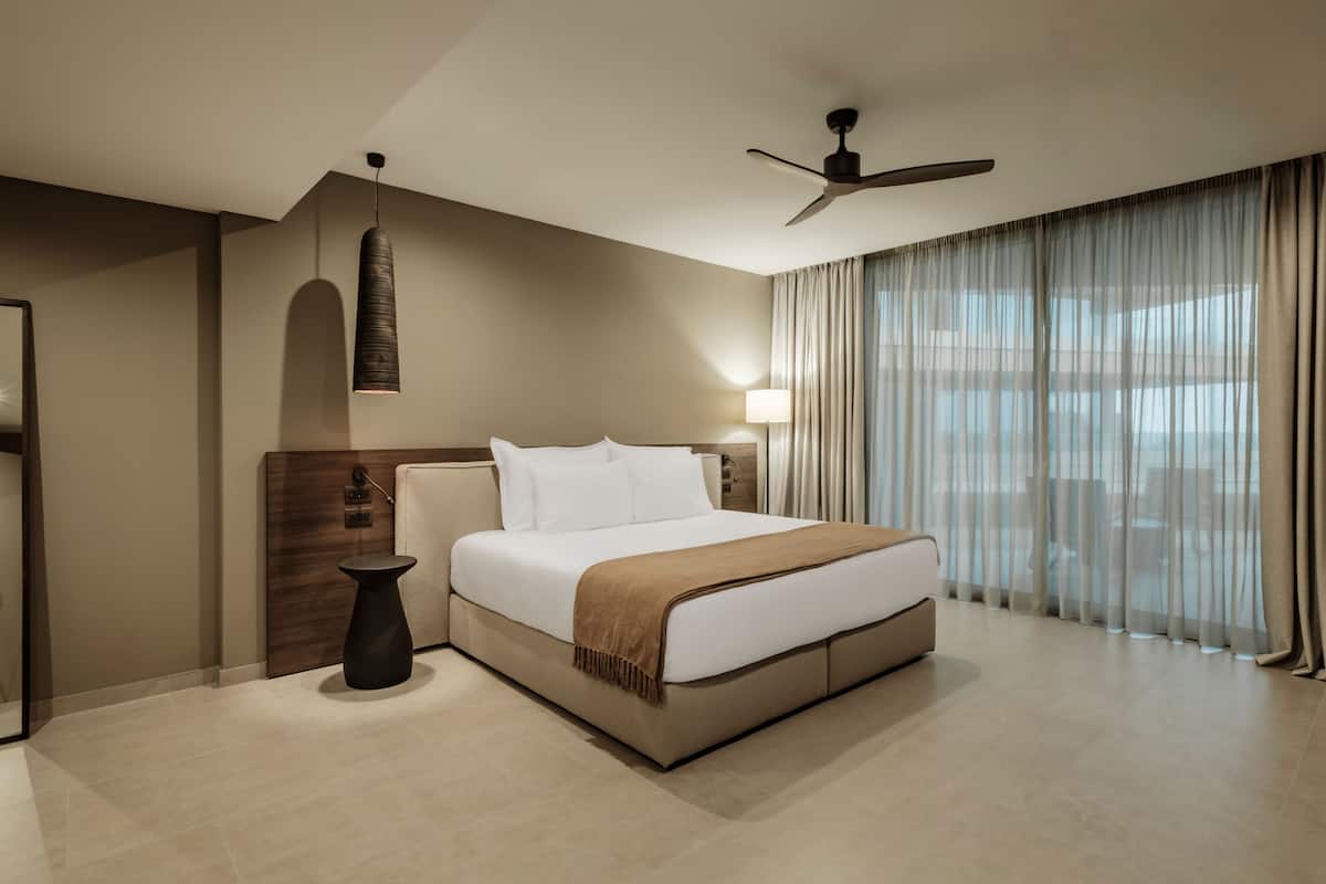 A modern hotel room with a large bed, a ceiling fan, a bedside lamp, and a view through sheer curtains.