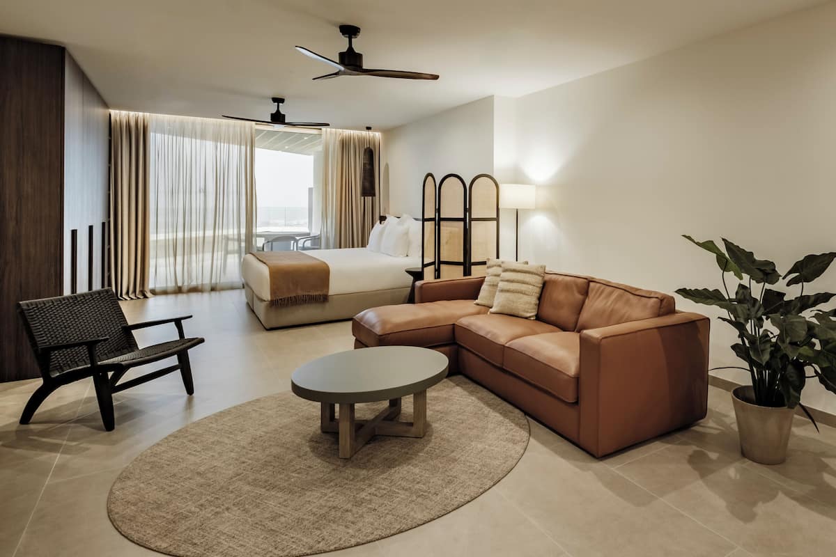A modern hotel room with a bed, a brown leather sofa, a round coffee table, a black chair, a potted plant, and a large window with curtains.