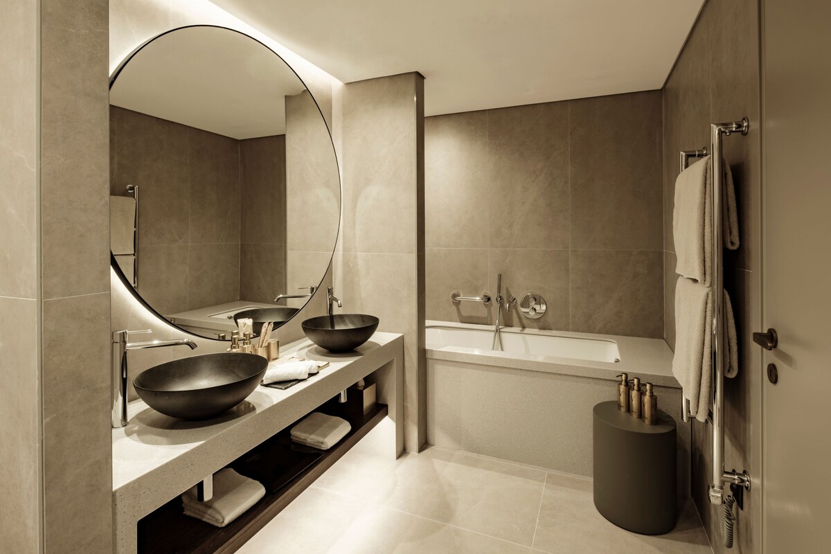 A modern bathroom with a large mirror, two sinks, a bathtub, and a round stool.