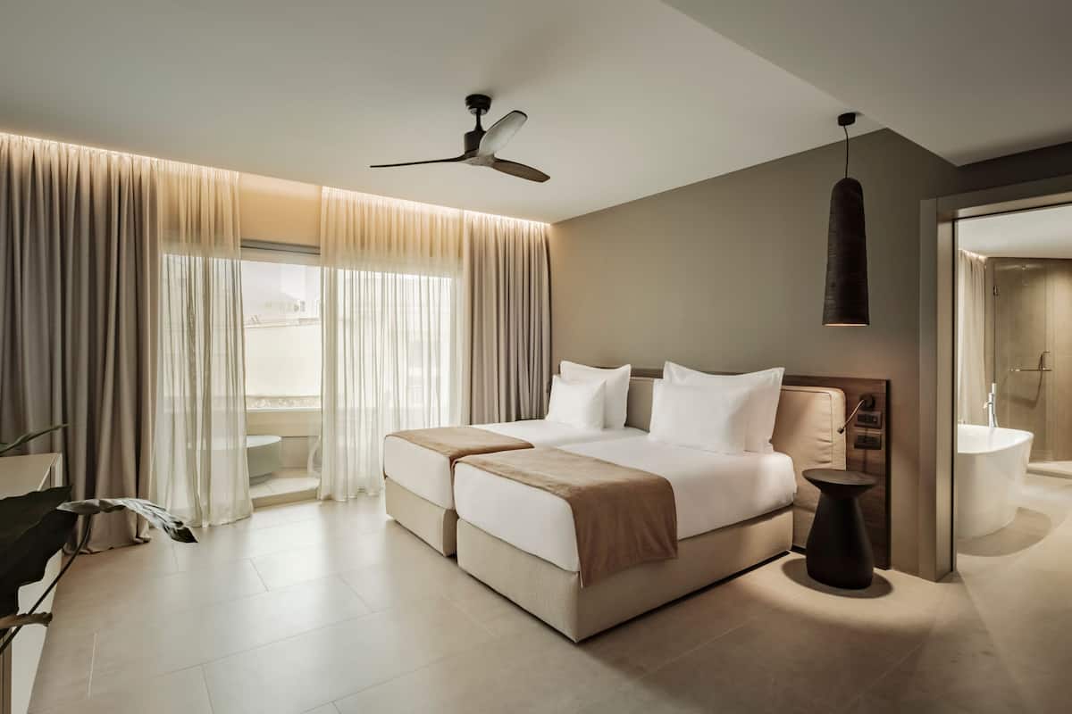 A modern hotel room with a large bed, a ceiling fan, a bathroom with a bathtub, and sheer curtains.