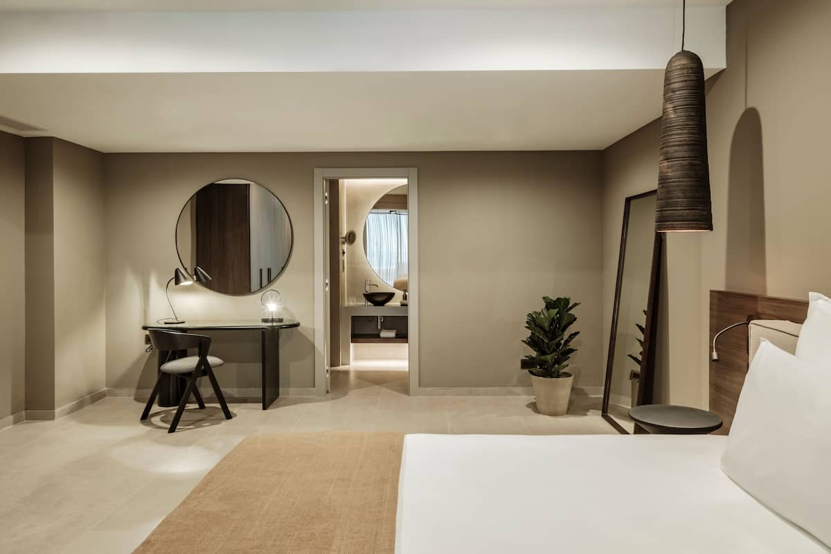A modern hotel room with a bed, desk, chair, mirror, potted plant, and a hanging light fixture.