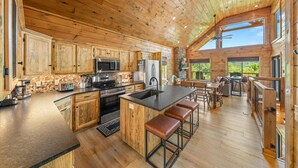 A spacious kitchen with wooden cabinets, a large island with stools, a stone accent wall, and a view of the outdoors.