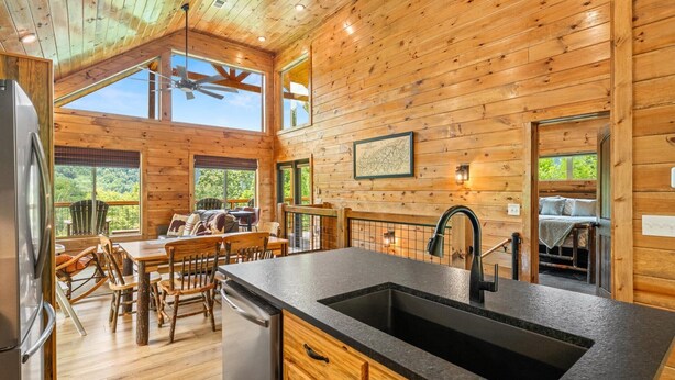 A wooden cabin interior with a kitchen, dining area, and a large window overlooking a forest.