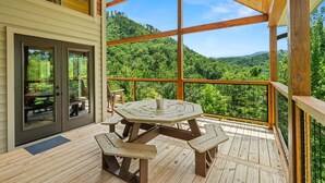 A wooden deck with a picnic table and benches, large windows offering a view of a forested landscape.