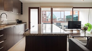 Una cocina moderna con armarios oscuros, encimera de granito y una isla grande. Cuenta con un comedor con mesa y sillas, y un amplio ventanal con vistas a la ciudad.