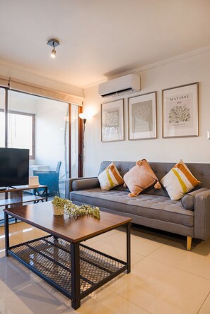 A modern living room with a grey sofa, a coffee table, and a TV. There are framed artworks on the wall and a window with blinds.