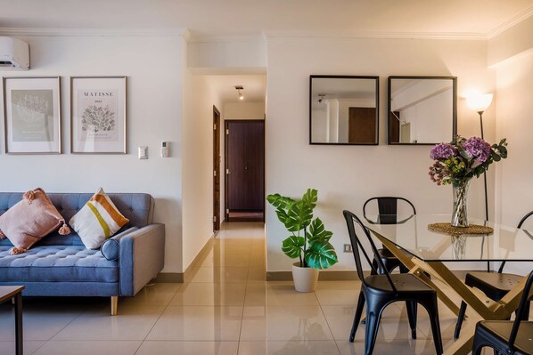 A modern living room with a blue sofa, two framed artworks, a dining area with a glass table and chairs, and a potted plant.
