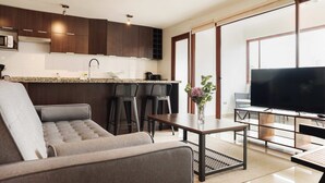 Una sala de estar moderna con sofá, una mesa de centro y televisor. Se observa una zona de cocina con armarios y microondas.