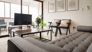 Una sala de estar moderna con un sofá, un televisor, una mesa de centro y dos sillones. Hay cuadros enmarcados en la pared y una planta sobre la mesa.