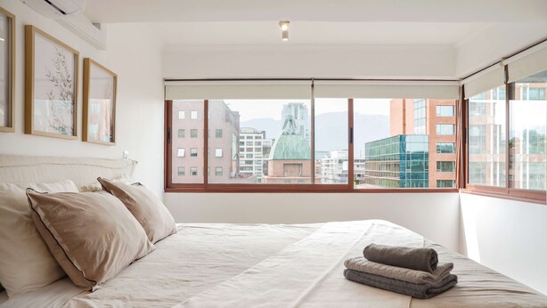 Un dormitorio con una cama grande, ropa de cama color crema y vistas a edificios de la ciudad a través de ventanales.