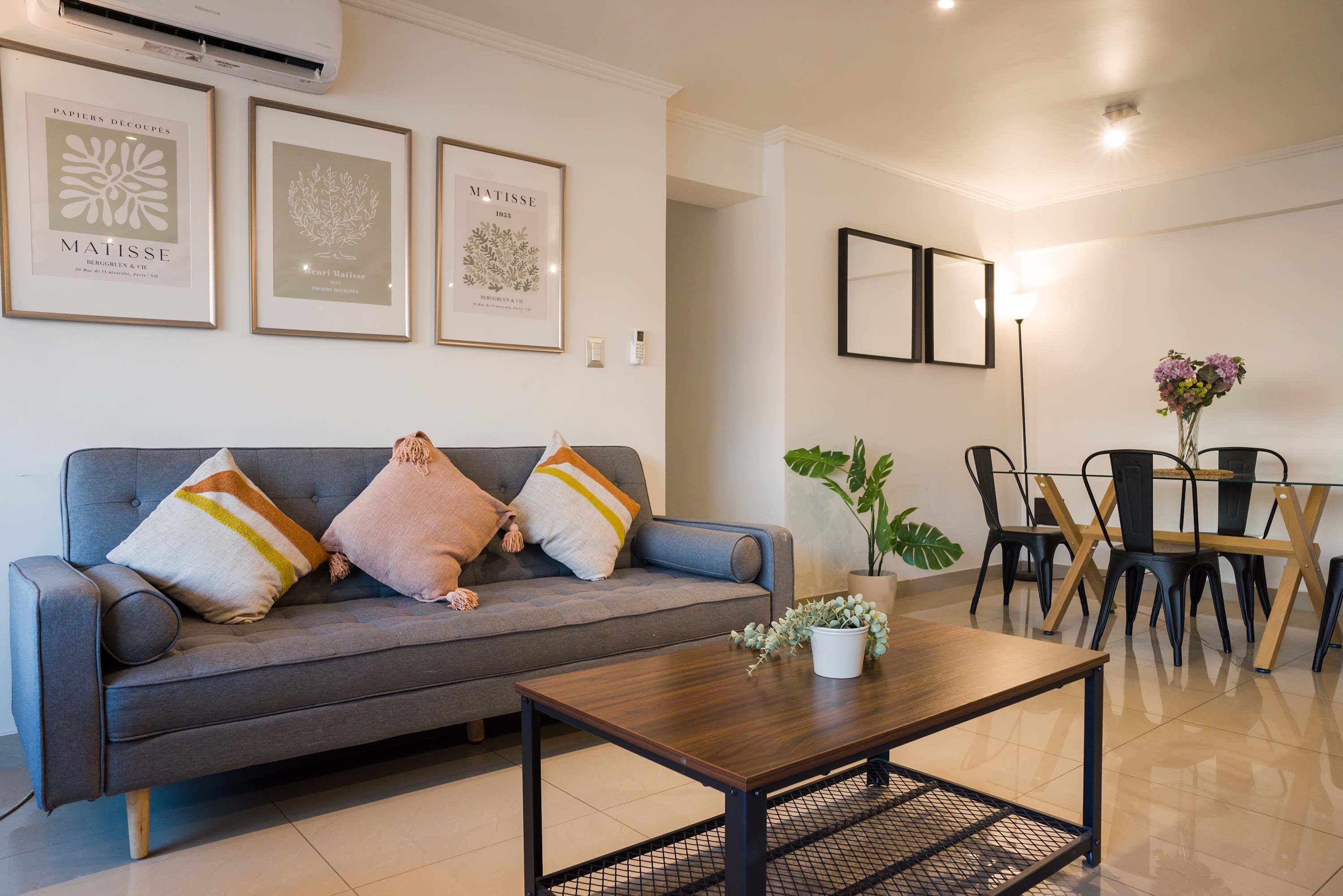 Uma sala de estar moderna com um sofá cinzento, uma mesa de centro e um espaço de jantar com mesa e cadeiras. Há quadros nas paredes e um vaso de planta na mesa de centro.